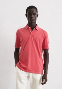Koraalkleurig polo shirt met een twee-knops sluiting en korte mouwen. Gemaakt van zachte stof met een royale pasvorm. Gecombineerd met lichtgekleurde broeken.