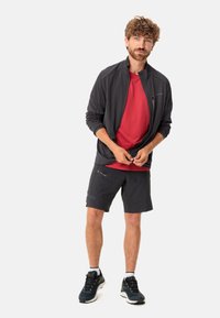 Mannen sportoutfit: zwarte zip-up jas, rode t-shirt, zwarte shorts en donkere schoenen met gele accenten. Glad materiaal, atletische pasvorm.