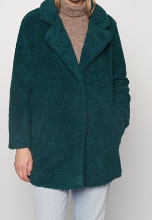 Tyrkis pelsjakke med en tekstureret, fluffy overflade, brede slag og sidelommer. Båret over en brun turtleneck og lyse jeans.