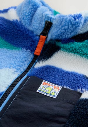 Fleece jacket with a blue, green, and white abstract pattern. Features a black pocket, orange zipper, and a patch logo. Soft texture.