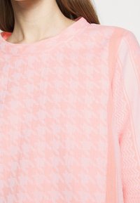 Jasnoróżowy sweter z wzorem w zygzak i teksturowanym materiałem. Dodatki obejmują wzorzystą cyfrę wzdłuż boku.