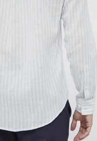 Camisa ligera a rayas en azul y blanco, con mangas largas y puños abotonados. Textura suave y detalle de dobladillo redondeado.