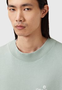 Sudadera verde claro con cuello acanalado y un pequeño detalle de logo blanco en el pecho. La tela parece suave con una textura lisa.