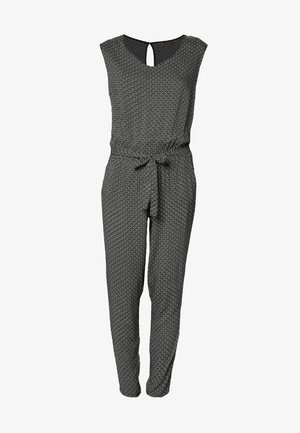 Vrouw draagt een zwart-witte jumpsuit met patroon en een gestrikte taille, een zwarte blazer, zwarte hakken en houdt een zwarte clutch vast.