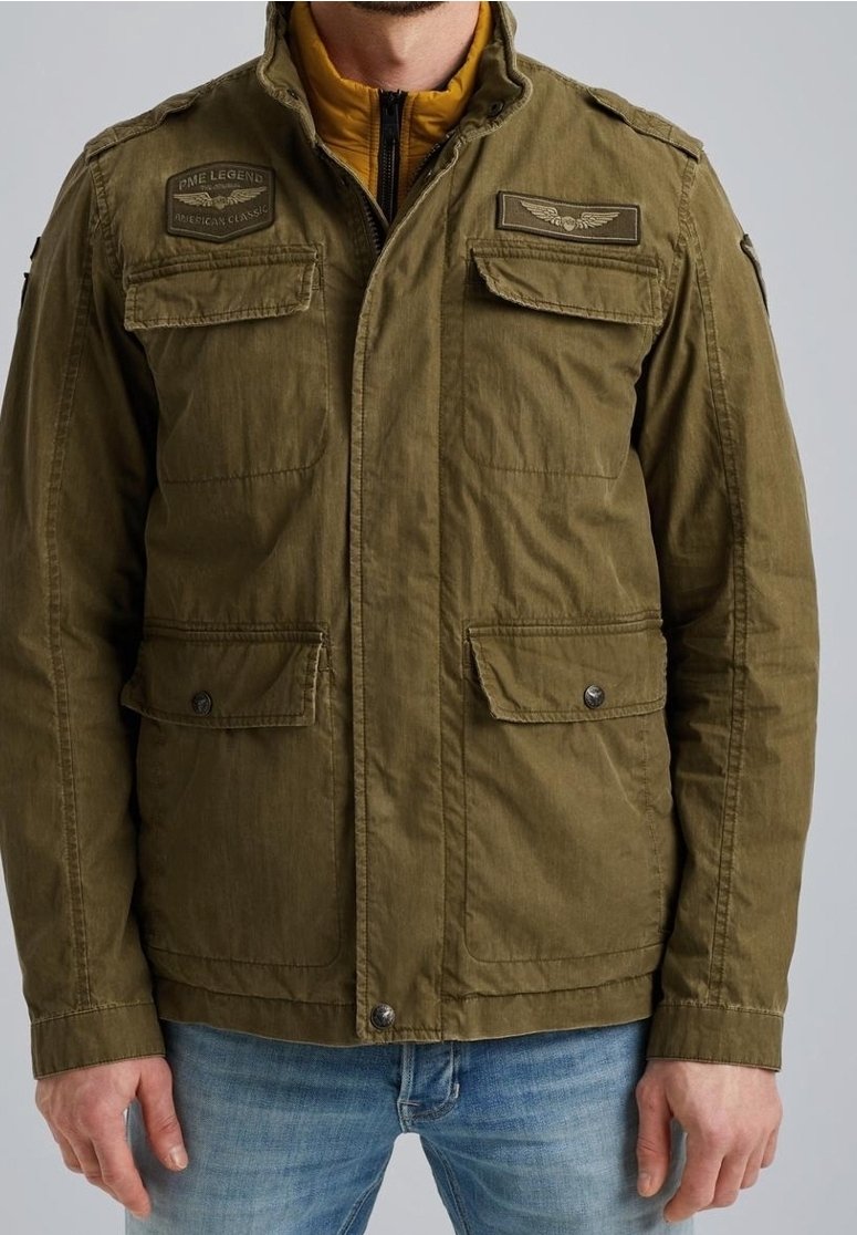 Homme portant une veste de style militaire vert olive avec des poches à rabat, des écussons sur la poitrine et la manche, par-dessus une couche zippée jaune moutarde.