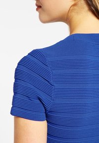 Modrý strukturovaný top s horizontálními žebrovanými pruhy, krátkými rukávy, kulatým výstřihem a hladkým materiálem. Zadní pohled ukazuje design ramene.
