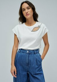 Hvid bomulds T-shirt med korte ærmer, med en udskæring og udsmykket bladdesign i sølv. Kombineret med højhalsede denimbukser.
