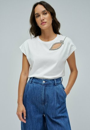 T-shirt en coton blanc avec des manches courtes, présentant une découpe et un design de feuilles rehaussé en argent. Associé à un pantalon en denim taille haute.