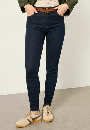 Mujer con jeans ajustados de color azul oscuro, cinturón de cuero marrón, chaqueta acolchada verde y zapatillas beige con rayas marrones, de pie sobre un suelo gris.