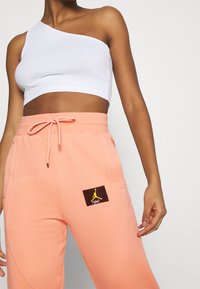 White ribbed crop top with one shoulder. Peach joggers featuring a Jordan logo patch, drawstring waist, and side pockets. Soft fabric texture.