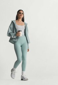 Lys teal vindjakke med hætte, grå tanktop, matchende teal leggings og hvide træningssko. Holder en kaffekop.