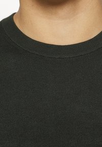 T-shirt a maniche corte verde scuro con collo a giro, caratterizzato da una finitura textured e cuciture sottili attorno al colletto. Visuale ravvicinata del tessuto.