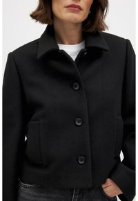 Chaqueta negra de mezcla de lana con un cuello estructurado, cierre frontal de botones y bolsillos laterales. Presenta una silueta corta y una textura suave.