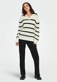 Pull rayé en crème et noir, doté d'un col haut avec une fermeture éclair. Le pull présente une texture de tricot épais et des manches amples. Assorti avec un pantalon noir.