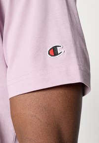 Helltaures Baumwoll-T-Shirt mit kurzen Ärmeln, das ein kleines schwarz-rotes gesticktes Logo am linken Ärmel aufweist. Weiche Textur.