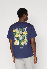 T-shirt em algodão azul marinho com um design floral, apresentando flores amarelas e letras cor-de-rosa a soletrar "JUVENTUS" nas costas. Mangas curtas.