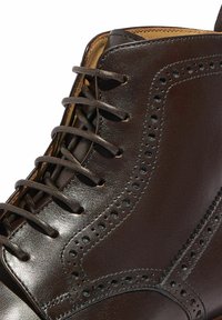 Botín de cuero marrón que presenta un acabado liso, detalles de brogue decorativos y una forma estructurada con cierre de cordones.