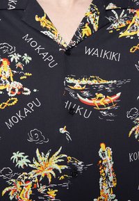 Chemise hawaïenne noire avec des scènes colorées d’îles, des palmiers, des pagayeurs en canoë, et des noms de lieux comme Waikiki et Mokapu.