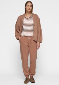 Losse bruine sweatshirt, gestreepte top met korte mouwen, bruine joggers met trekkoord en zachte bruine schoenen, tegen een neutrale achtergrond.