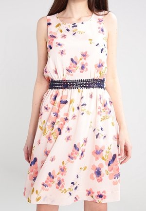 Femme portant une robe rose sans manches avec un imprimé floral bleu et pêche et une ceinture en crochet bleu marine à la taille.