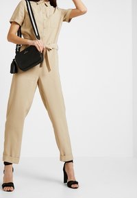 Combinaison beige à manches courtes, avec un boutonnage à l'avant et une ceinture à la taille. Associée à un sac bandoulière noir avec un pompon et une bandoulière rayée.