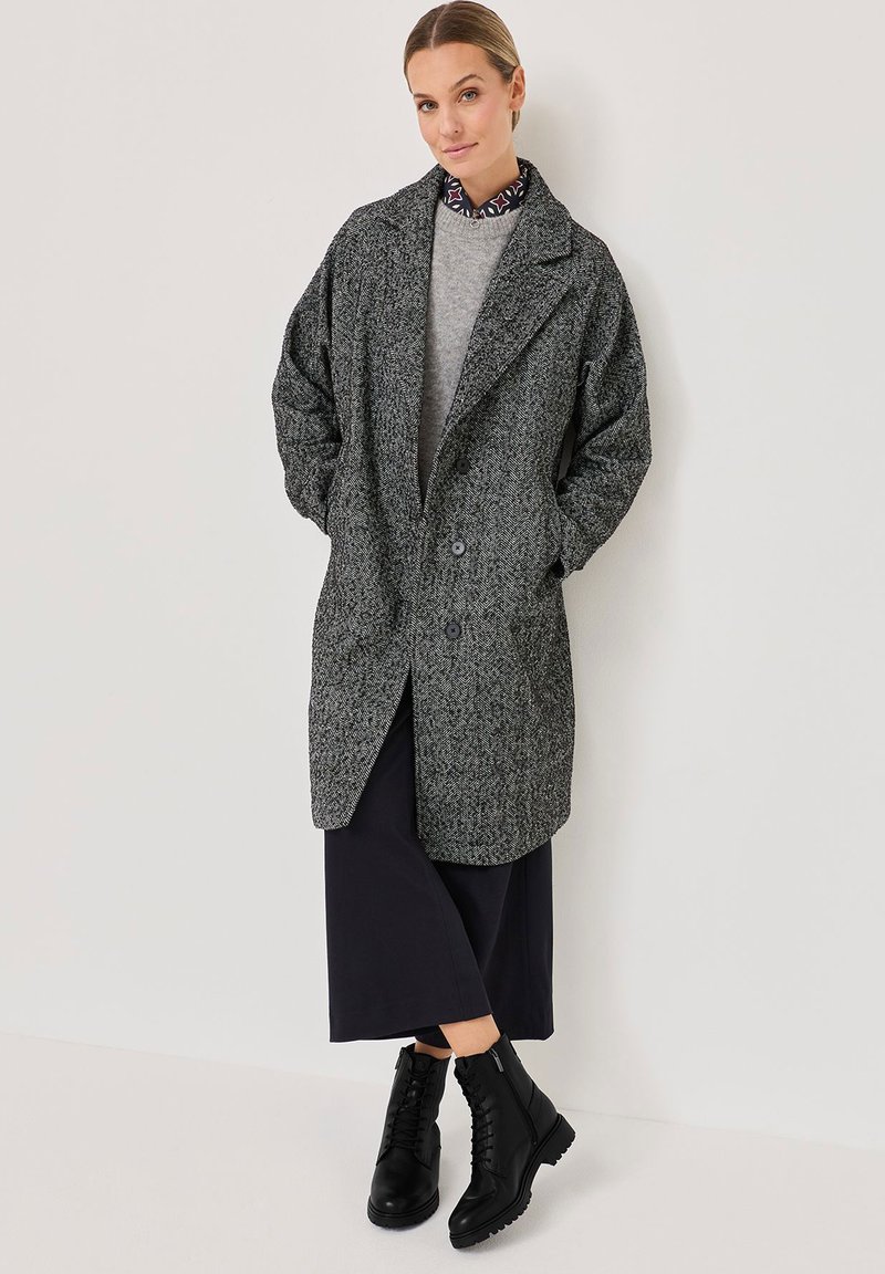 Manteau en pied de poule gris avec de grands revers, deux boutons et poches latérales, superposé sur un pull gris et une jupe noire, associé à des bottes noires.