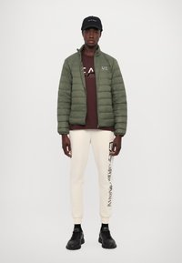 Padded green jacket, brown logo t-shirt, cream sweatpants with black text, and black sneakers. Features zip closure and ribbed cuffs.