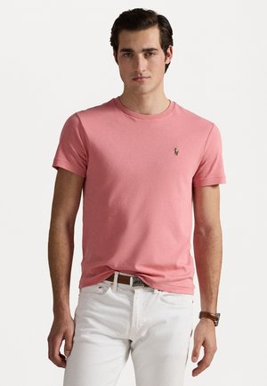 Korte mouwen roze katoenen t-shirt met een ronde hals en een klein geborduurd logo op de borst. Gdragen met witte broek en een bruine horloge.