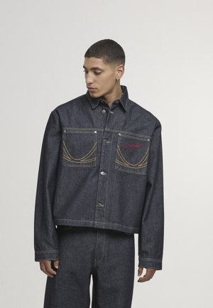 Tmavomodrá rifľová bunda s dvoma náprsnými vreckami, žltým prešitím a zapínaním na gombíky vpredu. Má špicatý golier a mierne skrátený strih.