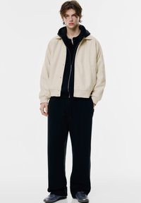 Beige uld-blend jakke med trykknapper over en sort hoodie og matchende sorte sweatpants, parret med mørke sneakers. Enkel design.