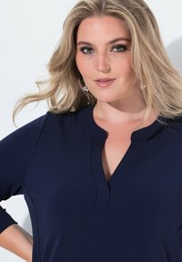Marineblaue Bluse mit rundem Halsausschnitt, Dreiviertelärmeln und einem dezenten V-Schlitz. Glattes Material mit leichtem Glanz.