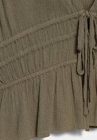 Gros plan sur un tissu vert olive froissé avec des coutures froncées et des liens en tissu noués sur le côté, montrant la texture et le détail du vêtement.