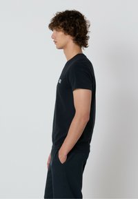 Čierne tričko s krátkym rukávom vyrobené z bavlny, s okrúhlym výstrihom a malým logom na hrudi, zobrazené na modeli zo strany.