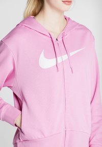 Sudadera con cremallera de color rosa hecha de tela suave, con un logo blanco de Nike en el frente. Incluye capucha y bolsillos laterales.