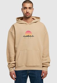 Beige hettegenser med kengurulomme, med et solnedgang- og bysilhuett-grafikk med teksten "MIAMI" under. Myk stoff, avslappet passform.