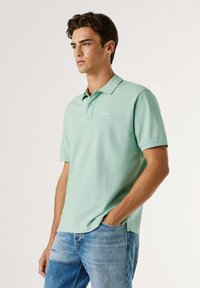 Ανοιχτοπράσινο πόλο από βαμβάκι, με γιακά, κοντά μανίκια και ένα μικρό λογότυπο στο στήθος, συνδυασμένο με ανοιχτό μπλε τζιν.