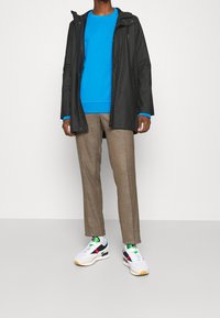 Chaqueta impermeable negra, sudadera azul, pantalones marrón claro y zapatillas multicolores con base blanca y suela amarilla, de pie frente a un fondo liso.
