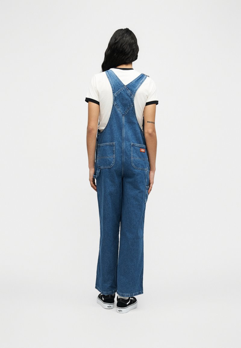 Donna con lunghi capelli neri che indossa una salopette di jeans blu e una maglietta bianca, vista di spalle su uno sfondo bianco uniforme.