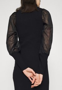 Femme portant une robe noire avec des manches bouffantes transparentes, vue de dos, les mains jointes derrière.
