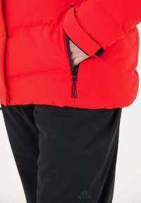 Czerwona ocieplana kurtka z kieszenią na zamek z czarnymi akcentami. Noszona na czarnych spodniach. Zbliżenie podkreśla strukturę i wzór.