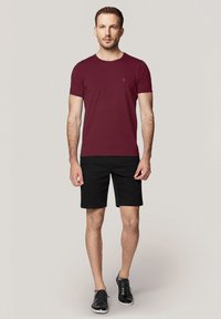 Bordowa koszula z krótkim rękawem i okrągłym dekoltem z małym logo, w połączeniu z czarnymi szortami i czarnymi butami, stojąca na neutralnym tle.