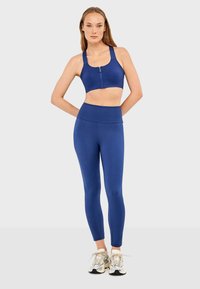 Brassière de sport bleue avec une fermeture éclair frontale et des bretelles larges, assortie à un legging taille haute, fabriquée dans un tissu lisse et extensible.