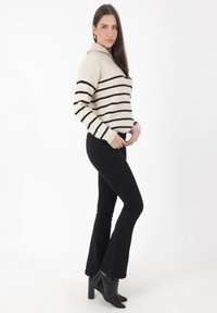 Cremfarbener und schwarzer gestreifter Strickpullover mit Kapuze, kombiniert mit schwarzen, ausgestellten Jeans und schwarzen, hochhackigen Stiefeletten, stehend vor einem weißen Hintergrund.