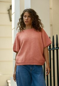 Lyserød strikket sweater med korte ærmer, afslappet pasform og ribkant, parret med blå vidde jeans. Den teksturerede stof er synlig.