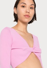 Camisola de manga longa rosa com decote em V profundo e design frontal torcido; o tecido parece suave com uma superfície ligeiramente texturada.
