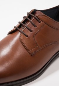 Chaussure habillée en cuir brun avec une texture lisse, des lacets droits et des détails cousus. Présente un bout rond et une semelle plate et foncée.