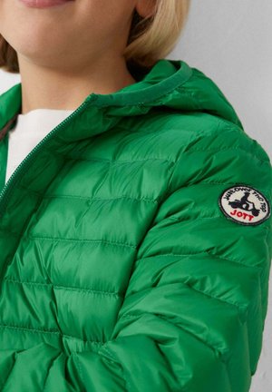 Primo piano di una persona che indossa una giacca trapuntata verde brillante con cappuccio e una toppa rotonda con il logo "JOTT" sulla parte superiore della manica.