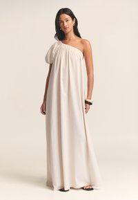 Abito lungo e fluente a spalla unica in tessuto beige chiaro con dettagli arricciati sul décolleté. Il modello indossa sandali neri e accessori.