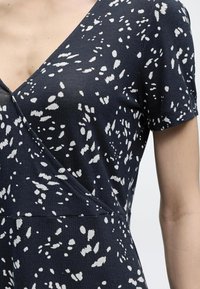 Κοντινό πλάνο ενός ατόμου που φοράει φόρεμα ναυτικομπλε με λευκά ακανόνιστα πουά σχέδια και κοντά μανίκια, εμφανίζοντας V-λαιμόκοψη και λεπτομέρειες ραφών στο πλάι.