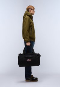 Černá taška typu duffle s nášivkou norské vlajky, držená osobou v olivově zelené bundě a modrých džínách, stojící na světlém pozadí.